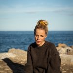 Bilde av Maria Sand sittende på et svaberg med sjøen bak.
